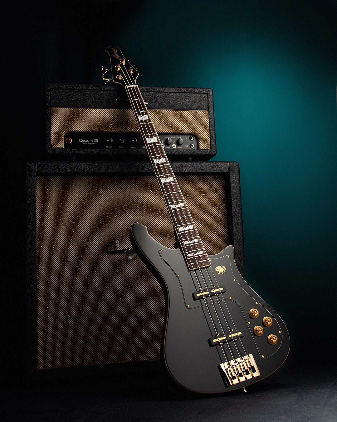 Black electric Baum nidhogg bass guitar with gold hardware in front of amplifiers on a dark background