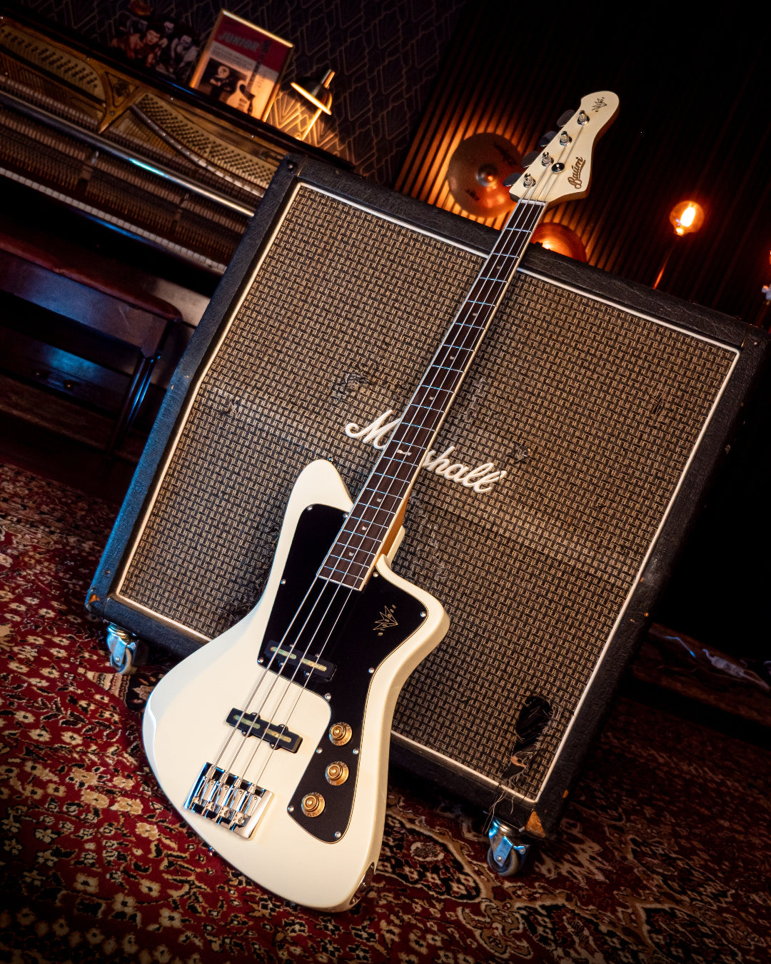 White Baum Wingman bass guitar leaning against a Marshall amplifier on a patterned rug.