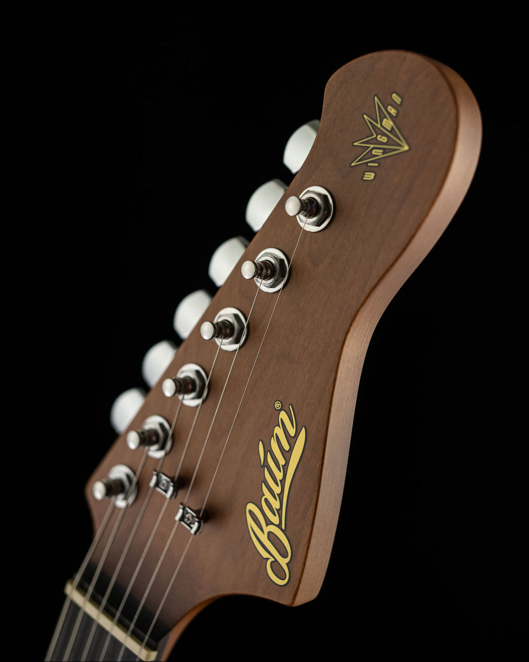 Close-up of a guitar headstock in dark brown wood with gold 'Baum' script and a gold crown logo, plus silver tuning pegs, shown against a black background.